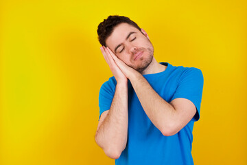 young handsome caucasian man wearing blue t-shirt against yellow background sleeping tired dreaming and posing with hands together while smiling with closed eyes.