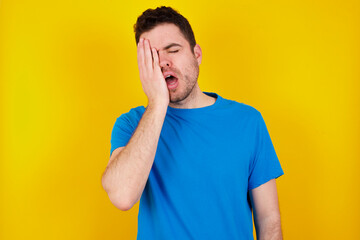 young handsome caucasian man wearing blue t-shirt against yellow background Yawning tired covering half face, eye and mouth with hand. Face hurts in pain.