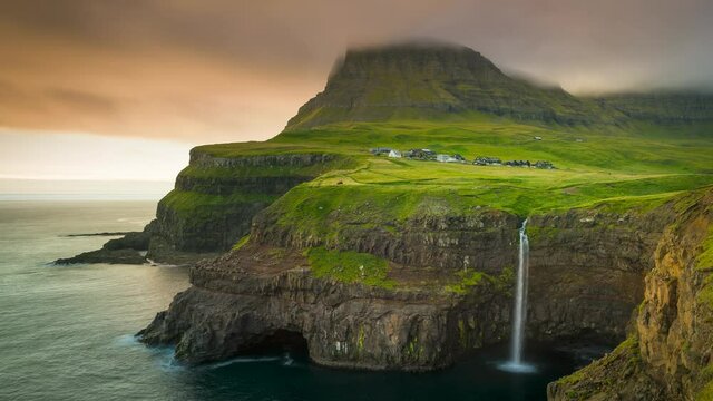 Time lapse of sunset over waterfall Mulafossur and village Gasadalur on Vagar, fast moving colourful clouds over mountain and ocean, Faroe Islands
