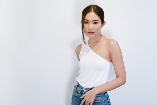 Portrait of fashionable beautiful young Asian woman in stylish strapless with jeans clothes with natural make-up and looking at camera standing near white wall. Lady slim wears white fit clothes.