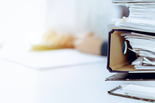 Binders with papers are waiting to be processed by business woman or bookkeeper working at the desk in office back in blur. Audit and tax concept