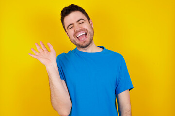 Overjoyed successful young handsome caucasian man wearing blue t-shirt against yellow background raises palm and closes eyes in joy being entertained by friends