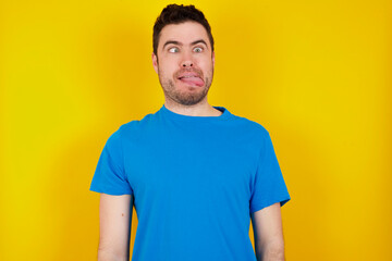 Funny young handsome caucasian man wearing blue t-shirt against yellow background makes grimace and crosses eyes plays fool has fun alone sticks out tongue.