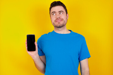 young handsome caucasian man wearing blue t-shirt against yellow background holds new mobile phone and looks mysterious aside shows blank display of modern cellular