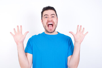 Optimistic young handsome caucasian man wearing blue t-shirt against white background raises palms from joy, happy to receive awesome present from someone, shouts loudly, Excited model screaming.