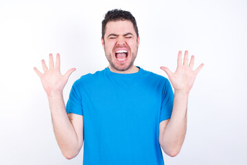 Fototapeta premium Emotive young handsome caucasian man wearing blue t-shirt against white background laughs loudly, hears funny joke or story, raises palms with satisfaction, being overjoyed amused by friend