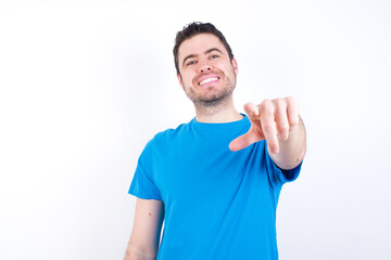 young handsome caucasian man wearing blue t-shirt against white background pointing at camera with a satisfied, confident, friendly smile, choosing you