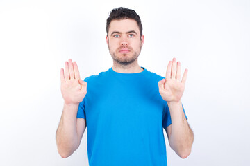 Serious young handsome caucasian man wearing blue t-shirt against white background pulls palms towards camera, makes stop gesture, asks to control your emotions and not be nervous