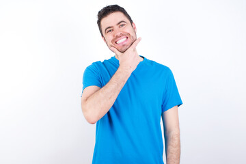 Obraz premium young handsome caucasian man wearing blue t-shirt against white background looking confident at the camera smiling with crossed arms and hand raised on chin. Thinking positive.