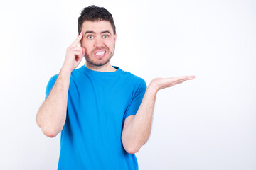 Fototapeta premium young handsome caucasian man wearing blue t-shirt against white background confused and annoyed with open palm showing copy space and pointing finger to forehead. Think about it.