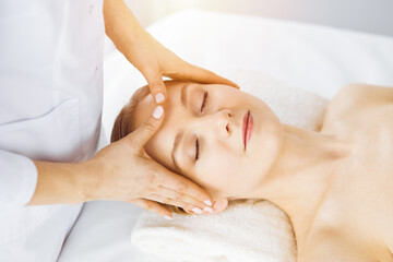 Fototapeta na wymiar Beautiful caucasian woman enjoying facial massage with closed eyes in sunny spa salon. Relaxing treatment in medicine