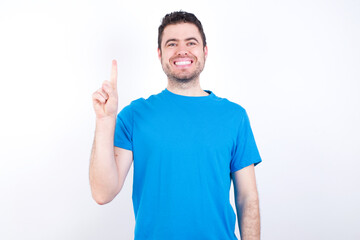 young handsome caucasian man wearing blue t-shirt against white background showing and pointing up with finger number one while smiling confident and happy.