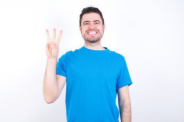 young handsome caucasian man wearing blue t-shirt against white background showing and pointing up with fingers number three while smiling confident and happy.