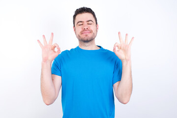 young handsome caucasian man wearing blue t-shirt against white background relax and smiling with eyes closed doing meditation gesture with fingers. Yoga concept.