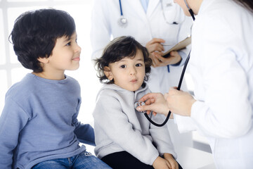 Woman-doctor examining a child patient by stethoscope. Cute arab toddler and his brother at physician appointment. Medicine concept