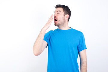 young handsome caucasian man wearing blue t-shirt against white background being tired and yawning after spending all day at work.