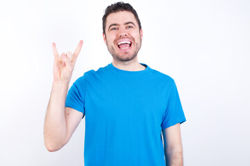 Fototapeta premium young handsome caucasian man wearing blue t-shirt against white background doing a rock gesture and smiling to the camera. Ready to go to her favorite band concert.