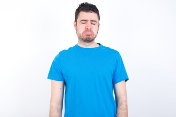 Obraz premium Dismal gloomy rejected young handsome caucasian man wearing blue t-shirt against white background has problems and difficulties, curves lower lip and closes eyes in despair, being in depression