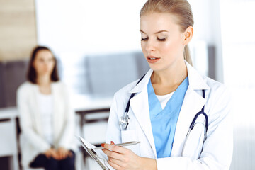 Woman - doctor is using clipboard for filling up medication history record form in clinic at the background of patient. Medicine concept