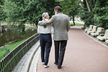 Back view of lovely mature couple walking in huggs at green park. Caucasian pensioners chatting on...