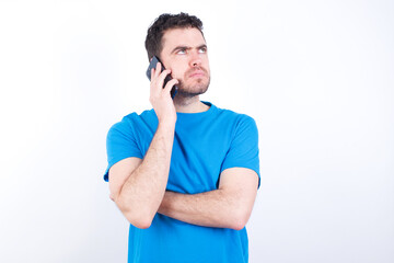 Sad young handsome caucasian man wearing blue t-shirt against white background talking on smartphone. Communication concept.