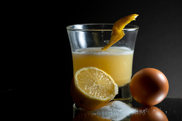 Whisky sour with egg white and lemon zest on the top of the glass. Single egg, half of lemon and sugar clod on the side. Reflecting surface and dark gradient background