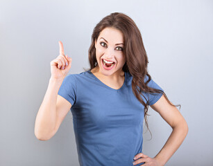 Fototapeta premium Beautiful excited casual woman with shock opened mouth pointing the finger up with smiling on empty copy space blue background. Closeup