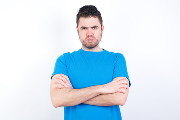 Gloomy dissatisfied young handsome caucasian man wearing white t-shirt against white background looks with miserable expression at camera from under forehead, makes unhappy grimace