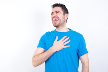 Joyful young handsome caucasian man wearing white t-shirt against white background expresses positive emotions recalls something funny keeps hand on chest and giggles happily.