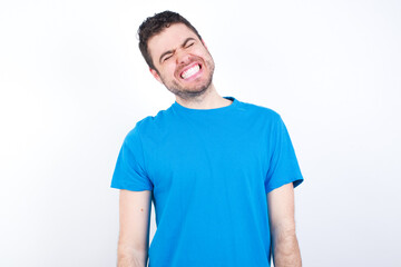 Fototapeta premium Positive young handsome caucasian man wearing white t-shirt against white background with overjoyed expression closes eyes and laughs shows white perfect teeth