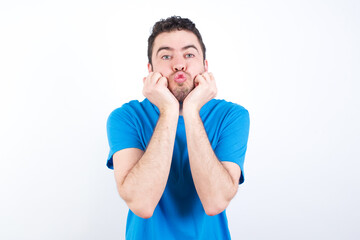 young handsome caucasian man wearing white t-shirt against white background with surprised expression keeps hands under chin keeps lips folded makes funny grimace