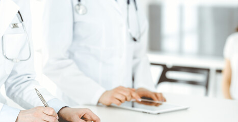 Unknown woman-doctors at work with patient at the background. Female physicians filling up medical documents or prescription while standing in hospital reception desk, close-up. Health care concept