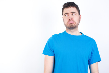 Fototapeta premium Dissatisfied young handsome caucasian man wearing white t-shirt against white background purses lips and has unhappy expression looks away stands offended. Depressed frustrated model.