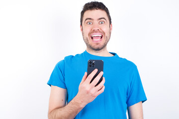 young handsome caucasian man wearing white t-shirt against white background holds mobile phone in hands and rejoices positive news, uses modern cellular