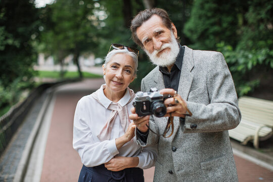 Caucasian elder couple in stylish outfits posing at green park with old fashioned photo camera in hands. Concept of family and retirement.