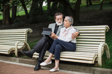 Beautiful senior couple in elegant clothes relaxing on wooden bench and using digital tablet. Happy family enjoying free time on fresh air with modern gadget.