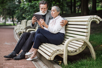 Fototapeta premium Happy mature couple using digital tablet while sitting in embrace on wooden bench. Relaxation at green park. Concept of modern technology.