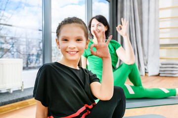 Sportive woman show okay gesture during fitness yoga exercises on mats near window, healthy family lifestyle. Focus on the girl.
