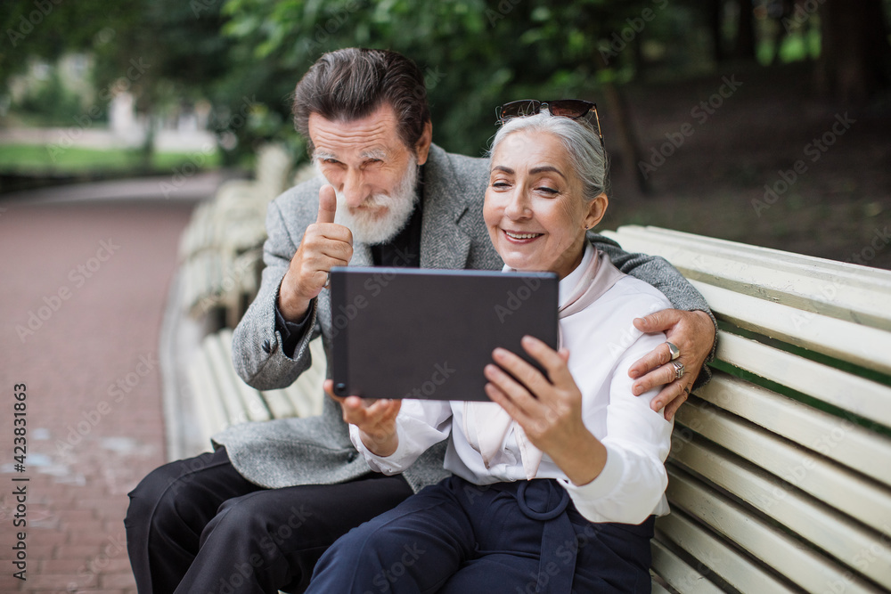 Wall mural Beautiful aged couple in stylish clothes having video conversation on digital tablet while relaxing at green park. Concept of people and technology.