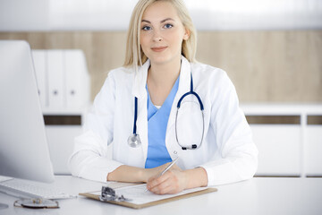 Portrate of woman-doctor at work while sitting at the desk in clinic. Blonde cheerful physician filling up medical form or prescription