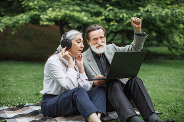 Cheerful aged man and woman using wireless laptop and headphones for listening music at green park. Senior couple spending time together with fun. - Powered by Adobe