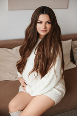 Beautiful pregnant female model with perfect makeup wearing white knitted dress holds her tummy and poses on the sofa