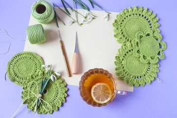 A cup of tea with a slice of lemon among handmade motifs of green color and needlework items on a blue background, top view