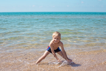 Little blond boy in swimming trunks is playing on the seashore with sand and water, smiling, standing in the water. Summer happy vacation.