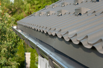 Gray metal roof tiles and rain gutter. House roofing system close-up on a green defocused background