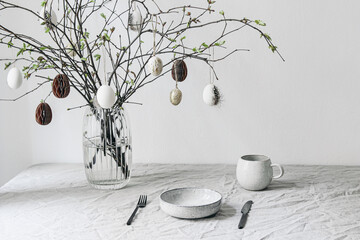 Easter festive table setting. Fresh branches in vase decorated by paper ornaments. Hanging chicken eggs with quail festher.Spring interior decoration. Scandinavian minimal design. Cup of coffee.