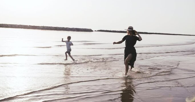 Side tracking shot of happy young mother having fun running along sunrise sea beach with little boy child slow motion.