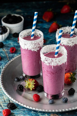 Berry smoothies with blue striped straws on a metal tray surrounded by various berries.