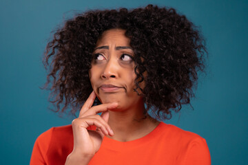 Fototapeta na wymiar Thinking mixed race woman looking up on blue studio background. Pensive face expressions. Pretty afro model with attractive appearance. 