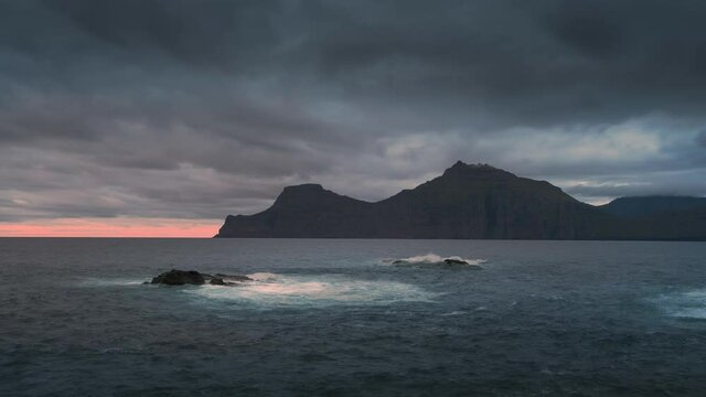 Time lapse with moving clouds at sunset over ocean and island Kalsoy, waves crashing againt rocks, Faroe Islands

